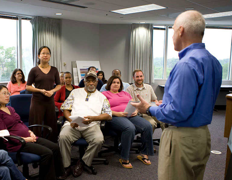 audience member stands among seated audience members who listen to a presenter standing at front of room