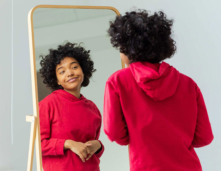 person in red sweatshirt looks at themself in full-length mirror