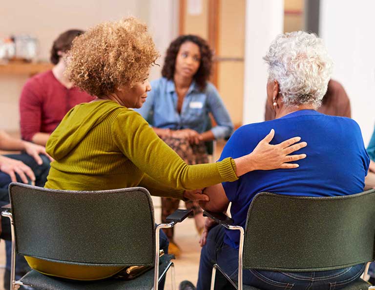 people sit in a circle of chairs and one person reaches over to place their hand on the back of the person next to them